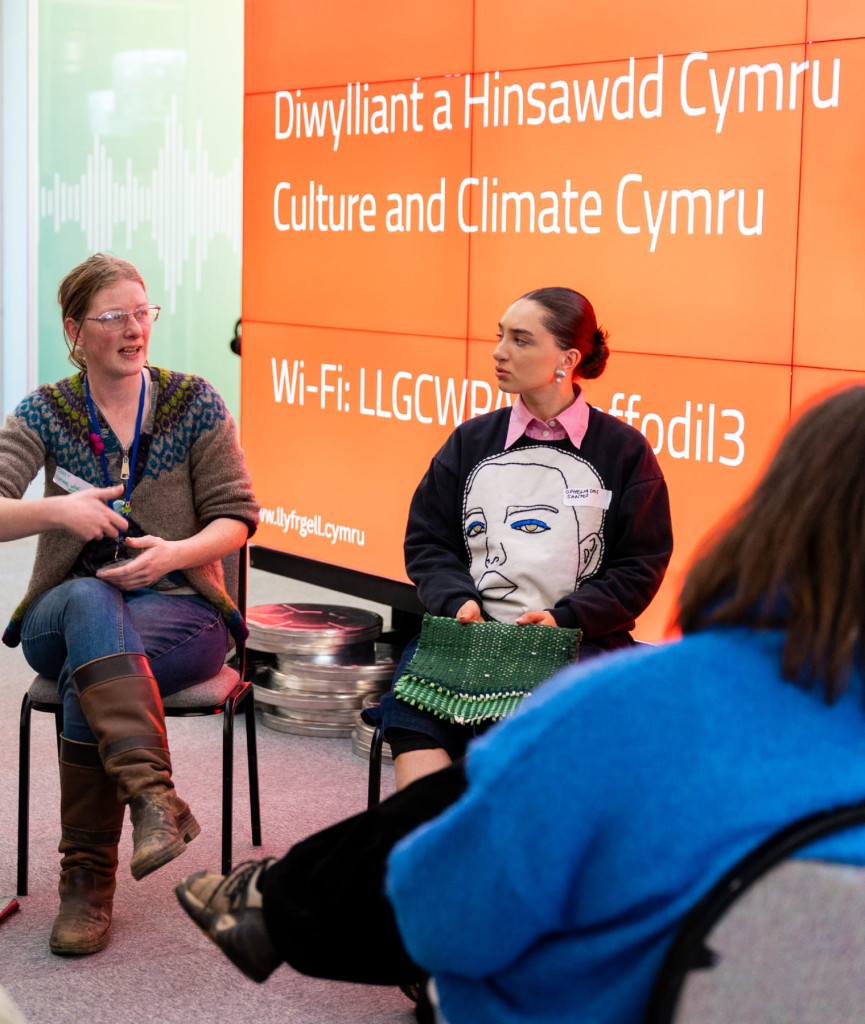 Mae dwy ddynes yn eistedd ac yn siarad o flaen wal oren sy'n arddangos manylion Diwylliant a Hinsawdd Cymru a Wi-Fi. Mae un ddynes yn ystumio tra bod y llall yn gwrando, gan ddal bag gwyrdd.